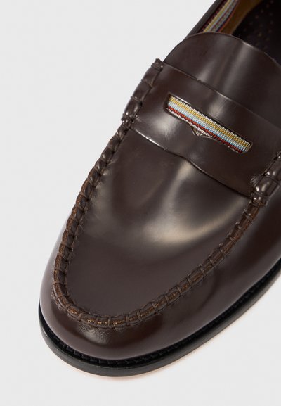 Mocassin en cuir marron avec coutures gravées et bande décorative aux rayures rouges, bleues et jaunes. Texture lisse et bout arrondi.