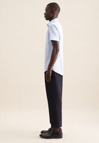 Camicia azzurra a maniche corte con colletto classico, abbinata a pantaloni sartoriali blu scuro e scarpe eleganti nere, vista di lato.