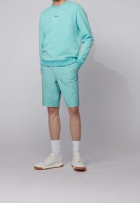 Uomo che indossa un maglione leggero blu e pantaloni corti abbinati, calzini bianchi e scarpe da ginnastica bianche, in piedi contro uno sfondo grigio.