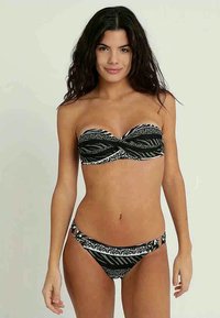 Ženska z dolgimi temnimi lasmi, oblečena v črno-belo bikinije z vzorcem brez naramnic, stoji ob svetli enobarvni podlagi, obrnjena proti kameri.