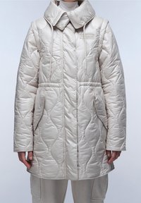 Béžová prošívaná bunda s vysokým límcem, stahovacími šňůrkami a dvěma předními kapsami. Materiál má lesklý povrch s vlnovým vzorem.