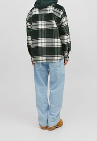 Grün-weißen Karomuster Flanellhemd, oversized Passform, mit abgerundetem Saum, kombiniert mit hellblauen Jeans und braunen Stiefeln.