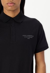 Juodas polo marškinėliai pagaminti iš medvilnės, su apykakle ir trimis mygtukais. Kairėje krūtinės pusėje yra iškilus logotipas.
