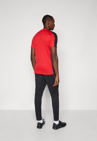 Rød sports T-shirt med sorte detaljer og en tætsiddende pasform, parret med sorte tapered bukser og sorte sneakers, set fra ryggen.