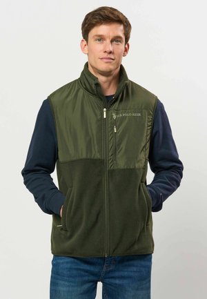Homme portant un gilet polaire vert foncé zippé sur des manches longues bleu marine, mains dans les poches, face à un fond clair uni.