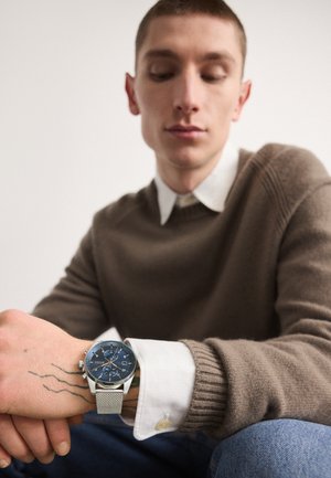 Sølvnet ur med en blå urskive, der har sølvfarvede detaljer, vist på en hånd med synlige tatoveringer. Brun sweater og hvid skjorte med krave er synlige.