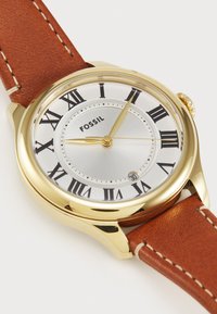 Montre-bracelet analogique dorée avec un cadran argenté, des chiffres romains et un bracelet en cuir brun ; présente un affichage de la date à la position 6 heures.