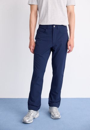 Person trägt marineblaue Hose, hellgraues Kurzarmhemd und silberne Turnschuhe und steht auf einem blauen Boden vor einer weißen Wand.