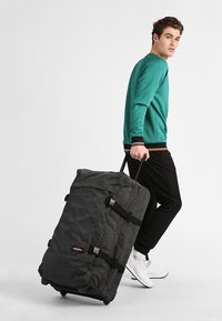 Bolsa de viaje de tela negra con ruedas, correas con hebillas y cremallera superior. Persona con sudadera verde y pantalones negros la arrastra.