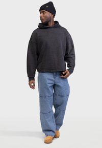 Felpa con cappuccio grigia scura dal taglio rilassato, abbinata a jeans larghi blu chiaro con dettagli di lavorazione a pezze e zoccoli marroni.