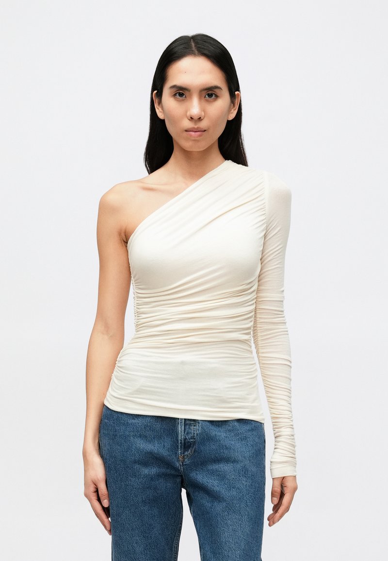 Top assimétrico de um ombro só em creme, com textura franzida e manga comprida ajustada, combinado com jeans de ganga azul.