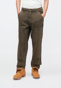 Φαρδιά παντελόνια χακί με εμφανείς ραφές, μπροστινές τσέπες και άνετη εφαρμογή, φορεμένα με ταμπά μποτάκια αστραγάλου με κορδόνια και λαστιχένια σόλα.