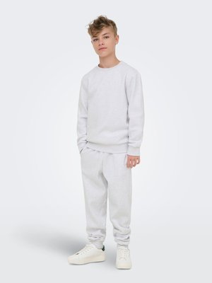 Boy with light brown hair wearing a light gray sweatshirt and matching sweatpants, standing with hands in pockets and white sneakers.