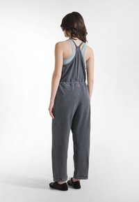 Mono de algodón gris con cintura ceñida y tirantes cruzados en la espalda. Piernas holgadas con puños enrollados, combinado con zapatos negros.