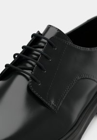Chaussure habillée en cuir noir avec une finition brillante, dotée de six œillets et de lacets noirs. La chaussure présente une texture lisse et des accents cousus.
