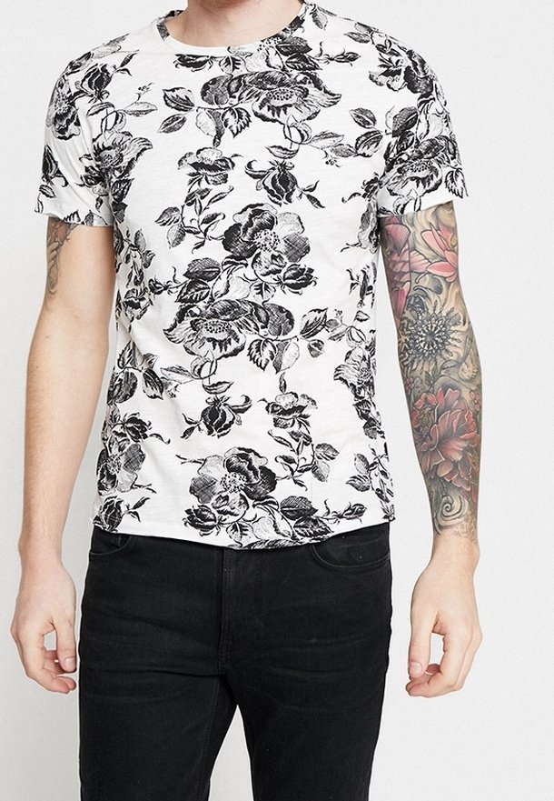 Homme portant un t-shirt blanc avec un imprimé floral noir et un pantalon noir, avec un tatouage floral coloré sur tout le bras droit.