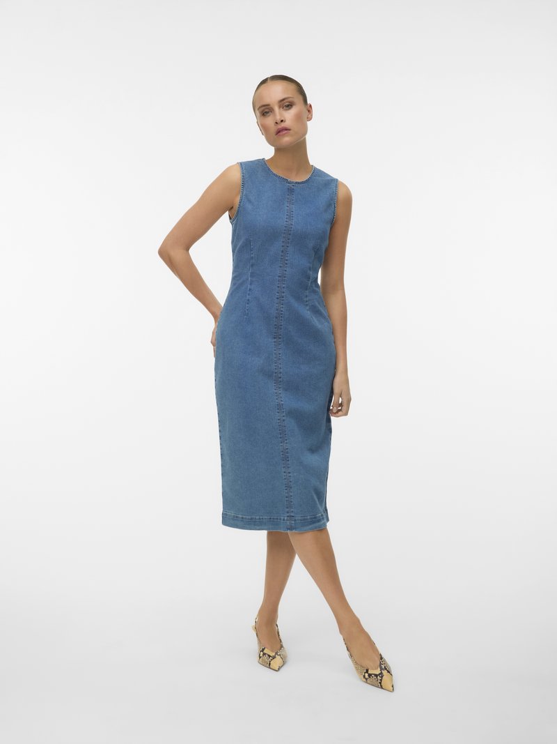 Femeie purtând o rochie albastră din denim fără mâneci, până la genunchi, și pantofi cu toc ascuțit, print piele de șarpe, stând cu un braț pe spate.