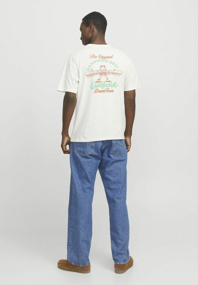 Hvid bomulds t-shirt med et farverigt grafisk design på ryggen med tekst. Bæres med blå denim jeans og brunt fodtøj.