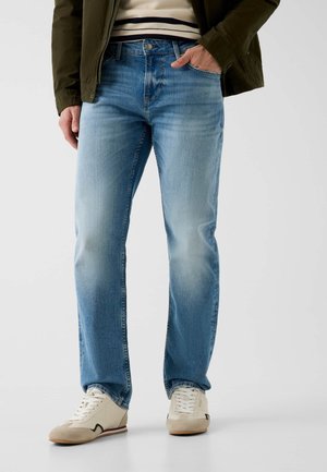 Mann trägt hellblaue Jeans, weiße Sneakers, gestreiftes Hemd und dunkelgrüne Jacke, steht mit einer Hand in der Tasche vor einfarbigem Hintergrund.