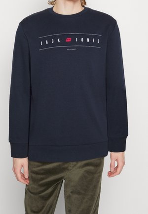 Osoba nosząca granatową bluzę z napisem i logo "JACK JONES" na piersi, w połączeniu z oliwkowozielonymi sztruksowymi spodniami, stojąca na białym tle.