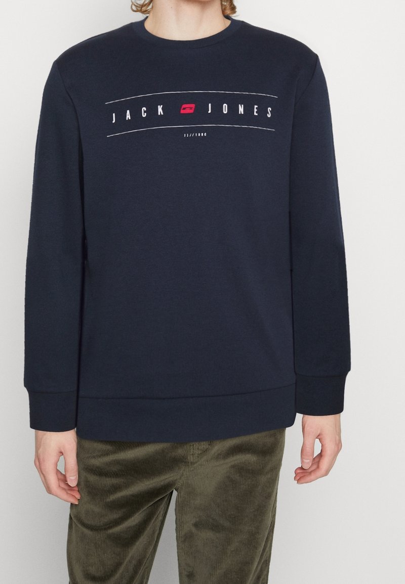 Personne portant un sweat-shirt bleu marine avec le texte et le logo "JACK JONES" sur la poitrine, assorti d'un pantalon en velours côtelé vert olive, debout devant un fond blanc.