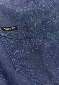 Jeanskjorte i mørkeblå med et struktureret mønster, med et sort "GUESS" label på lommen og en klassisk skjortekrave.
