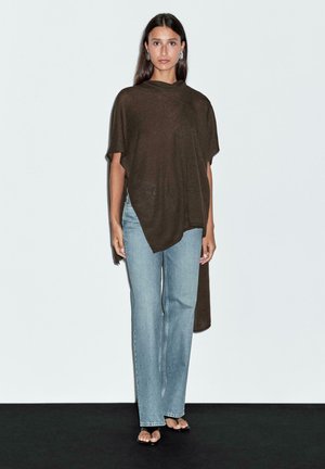Brauner übergroßer Poncho mit drapiertem Design, kurzen Ärmeln und längerem Rückensaum, kombiniert mit hellblauen, gerade geschnittenen Jeans.