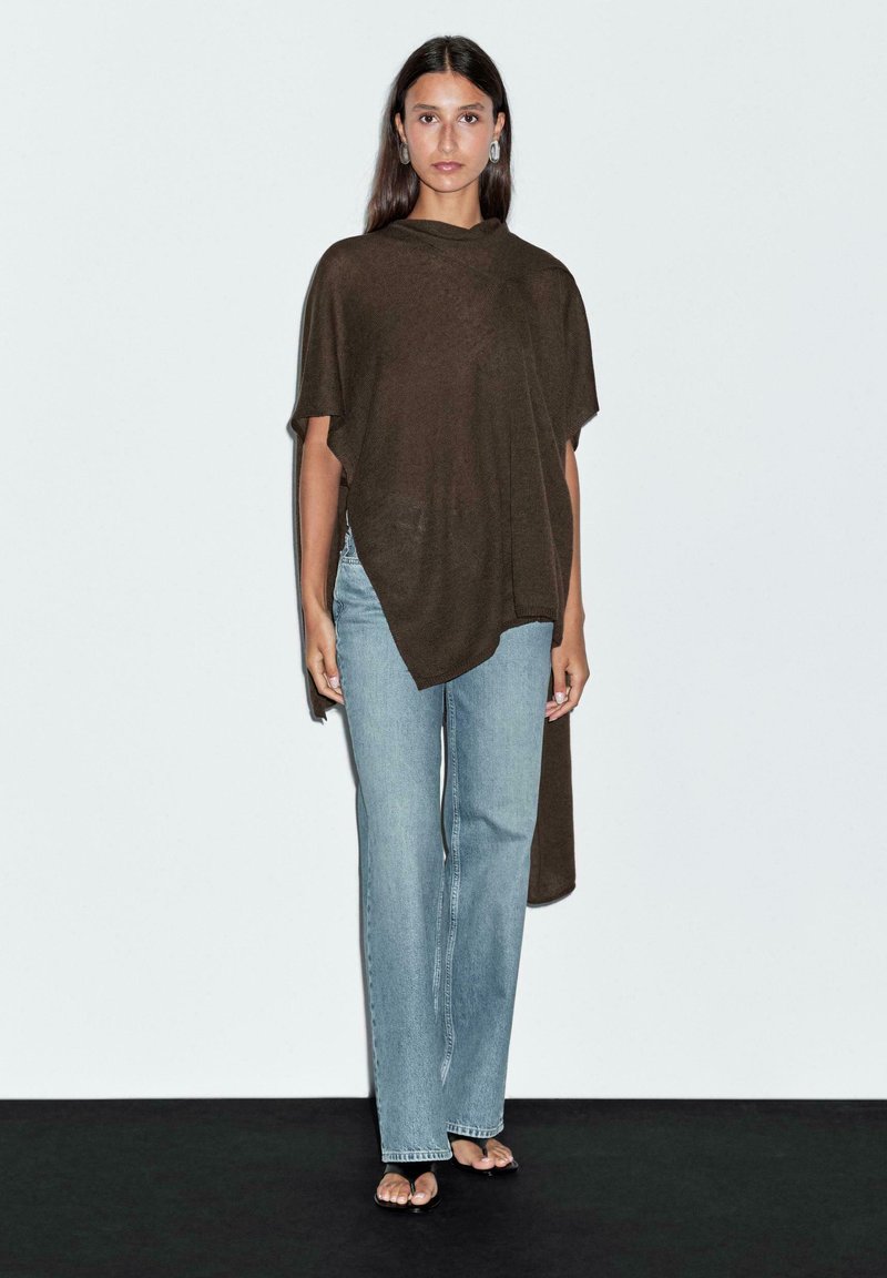 Brun oversized poncho med draperad design, korta ärmar och längre bakstycke, i kombination med ljusblå raka jeans.