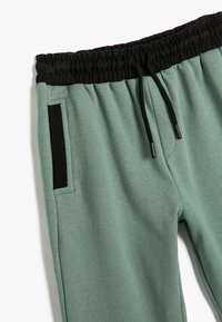 Grüne Sweatpants mit einem schwarzen elastischen Bund, verstellbarem Kordelzug und einer Seitentasche mit einem kontrastierenden schwarzen Streifen.