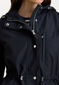 Námorně modrá lehká bunda se zipem vpředu, patentkami, stahovací šňůrou v pase a kapucí. Obsahuje náprsní kapsu a stříbrné kovové doplňky.