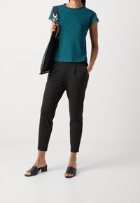 Donna che indossa un top corto di colore teal, pantaloni neri alla caviglia, mule aperte nere con tacco e una borsa nera con tracolla a catena.