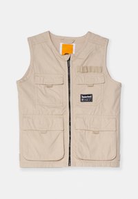 Beige arbetsväst med blixtlås framtill, flera stora fickor och en logotypmärke, med en mjuk textur och ärmlös design.