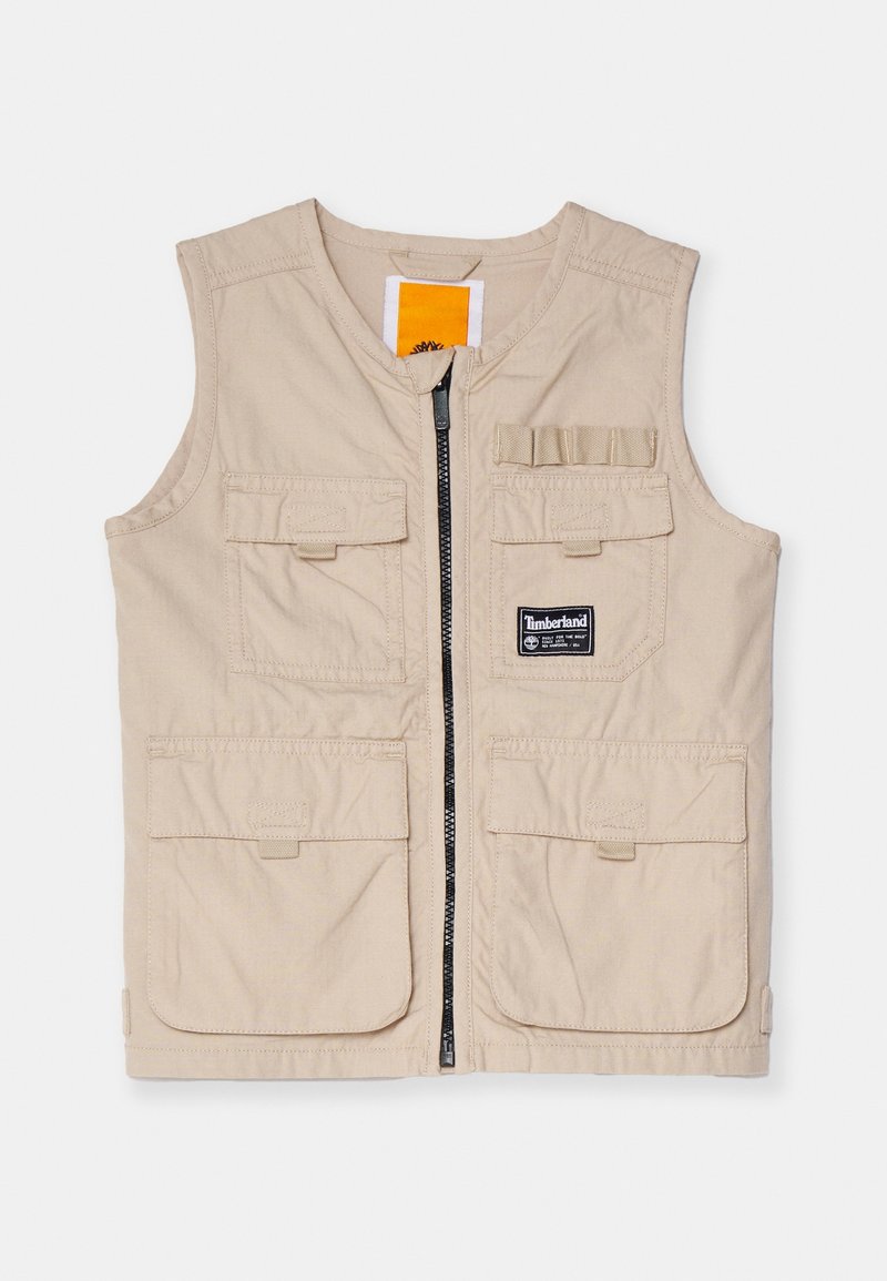 Beige arbetsväst med blixtlås framtill, flera stora fickor och en logotypmärke, med en mjuk textur och ärmlös design.