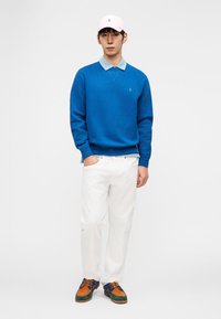 Άνδρας που φοράει μπλε φούτερ, ανοιχτό μπλε πουκάμισο με γιακά, λευκό παντελόνι, πολύχρωμα παπούτσια και ανοιχτό ροζ καπέλο μπέιζμπολ, στέκεται μπροστά σε λευκό φόντο.