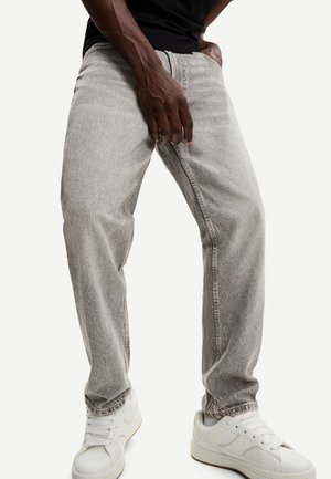 Homme portant un jean gris clair ample, une chemise noire et des baskets blanches, debout avec une main dans la poche devant un fond uni.