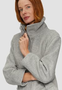 Hellgraue, im Fischgrätmuster gehaltene Jacke mit hohem Kragen, ausgestattet mit einem Reißverschluss und Druckknopfdetails am Kragen.