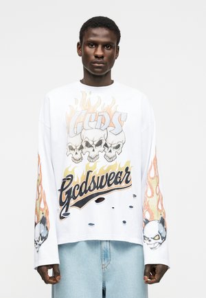 Jaunas vyras dėvi baltą ilgomis rankovėmis marškinius su liepsnojančiomis kaukolėmis ir užrašu „Gcds“, derinamus su šviesiai mėlynomis džinsais.