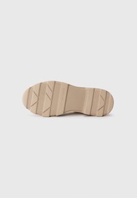 Semelle d'une chaussure, en caoutchouc beige avec une surface texturée, présentant des rainures horizontales et une zone de marque. Fond plat pour l'adhérence.