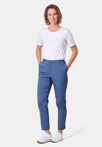 Pantalons bleus ajustés avec plis sur le devant, fermeture éclair et bouton, associés à une chemise blanche à manches courtes et des baskets blanches.