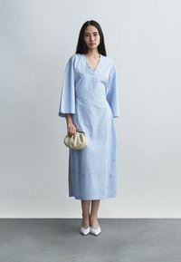 Γυναίκα που φοράει ένα ανοιχτό μπλε φόρεμα με μακριά μανίκια και μήκος μέχρι τους αστραγάλους, με διακριτικά σχέδια, κρατώντας μια μπεζ τσάντα φάκελο, και στέκεται μπροστά σε απλό φόντο.