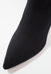 Gros plan sur une chaussure noire en tissu tricoté à bout pointu couvrant l'avant du pied sur un fond blanc.
