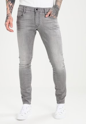 Slim fit lysegrå jeans båret af en tatoveret person, der står med den ene hånd i lommen og hvide sneakers.