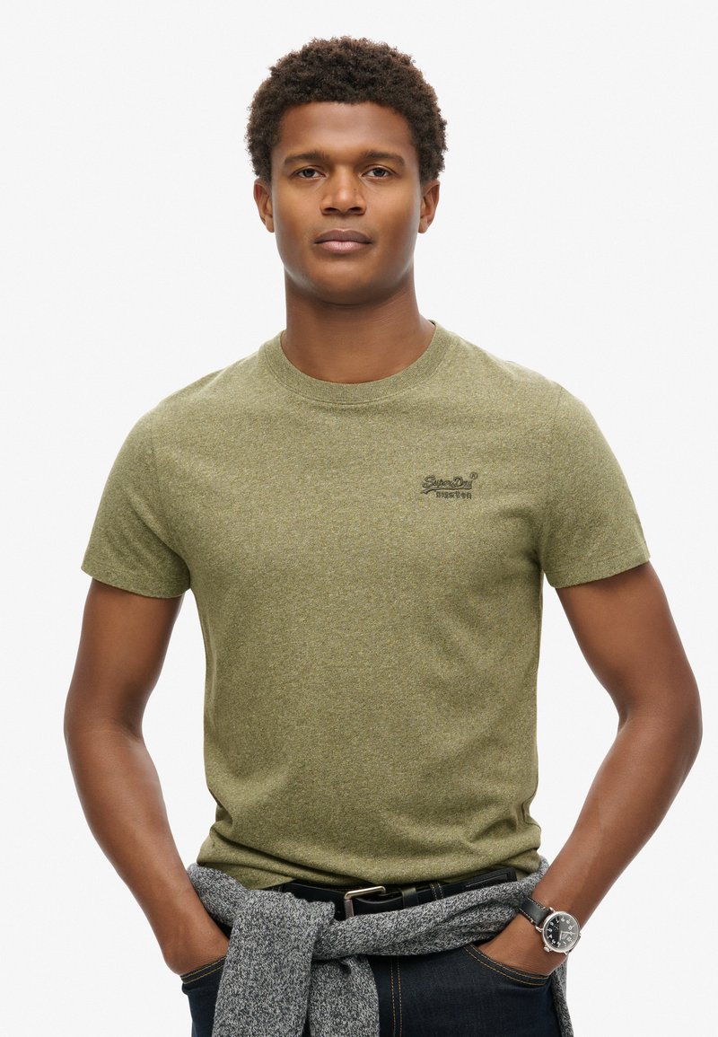T-shirt verde in cotone tessurizzato con collo rotondo e maniche corte. Presenta un piccolo logo sul petto. Jeans in denim scuro e maglione grigio annodato in vita.