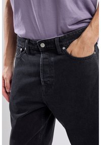 Mustat farkut, joissa on suora leikkaus, etutaskut, metallinappisuljenta ja huomaamattomia ompeluyksityiskohtia. Yhdistetty vaaleanpurahein paidan kanssa.