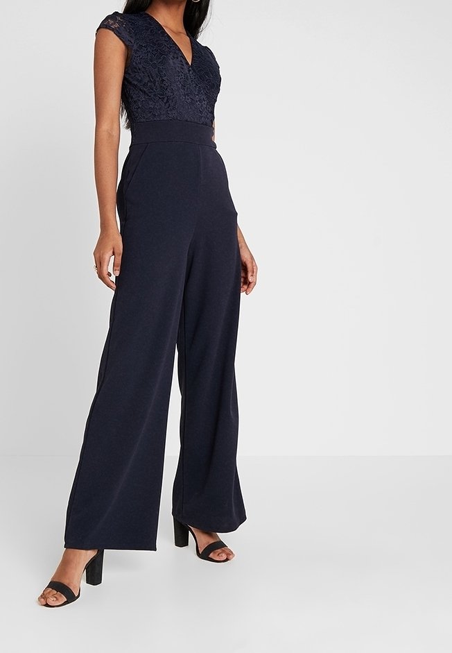 Combinaison bleu marine avec un corsage en dentelle, des manches courtes, un décolleté en V profond et un pantalon large. Portée avec des sandales à talons noires.
