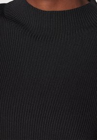 Gros plan d'une personne portant un pull noir côtelé à col montant, montrant la texture et le détail du col.