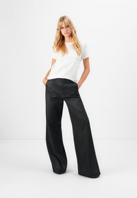 Donna bionda che indossa occhiali trasparenti, top bianco a maniche corte e pantaloni neri a gamba larga, in piedi con le mani nelle tasche su uno sfondo bianco.
