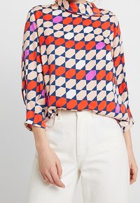 Blouse à motifs géométriques colorés avec des tons rouges, bleus et beige, dotée d'un col chemise et de manches trois-quarts. Texture de tissu lisse.