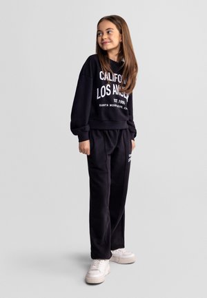 Młoda dziewczyna stojąca, ubrana w czarną bluzę i spodnie z białym napisem, białe sportowe buty, uśmiechnięta i patrząca w lewo.