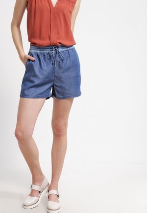 Vrouw draagt een mouwloze blouse in roestkleur, blauwe denimshorts met elastische taille en trekkoord, en witte Mary Jane-schoenen, staand.